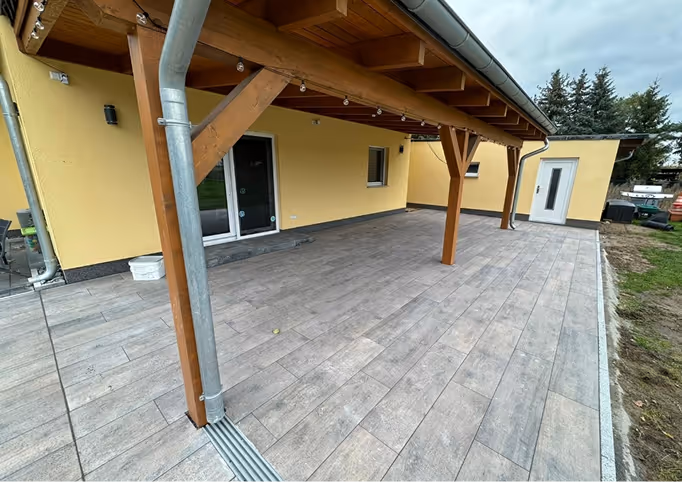 Überdachte Terrasse mit hellgrauen Fliesen, Holzbalken und gelber Hausfassade unter bewölktem Himmel.