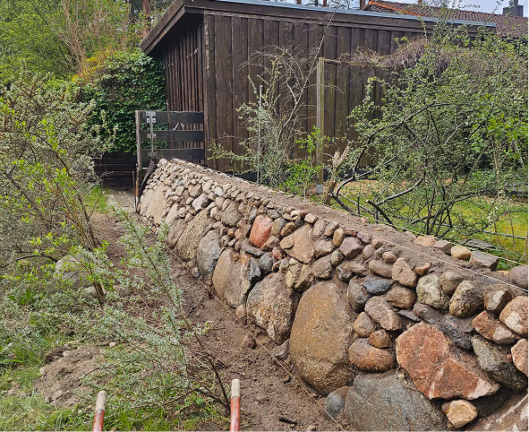 Natursteinmauer entlang eines Gartens mit grünem Buschwerk und einem Holzgartenhaus im Hintergrund.