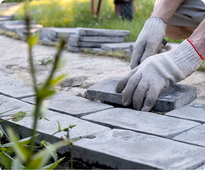 Hände mit Arbeitshandschuhen legen graue Pflastersteine auf einen Sanduntergrund im Garten.