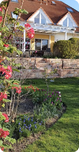 Ein Haus mit rotem Dach, Terrasse mit Pergola, davor blühende Blumen und gepflegter Rasen.