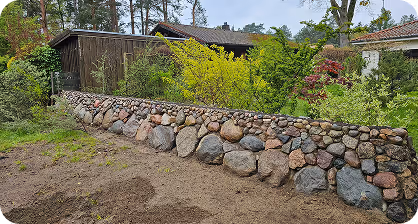 Steinerne Trockenmauer mit bunten Steinen vor grünen Büschen und Holzhäusern.