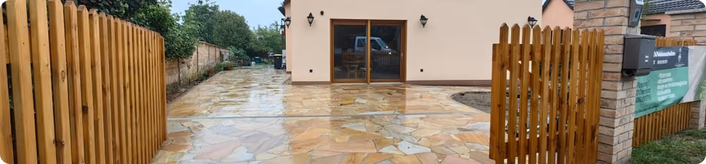 Offener Holzzaun mit Natursteinplatten als Hof vor einem modernen Haus mit Außentüren aus Glas.