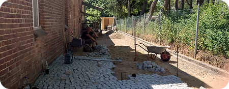 Arbeiter verlegen Pflastersteine entlang einer Backsteinmauer mit Schubkarre und Werkzeug.