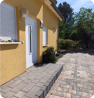 Haus mit gelber Fassade, weißer Eingangstür und gepflastertem Gehweg unter blauem Himmel.