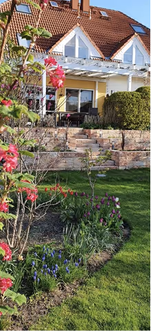 Haus mit rotem Ziegeldach und Terrasse mit Glasdach, davor blühender Garten mit bunten Blumen und grünem Rasen.
