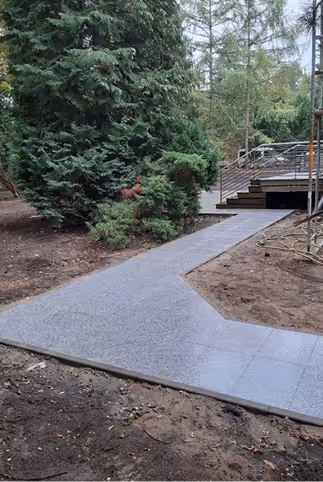 Gepflasterter Gehweg, der sich durch einen Garten mit großen Tannenbäumen und brauner Erde zum Hauseingang schlängelt.