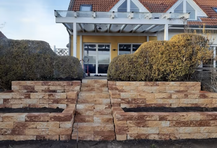 Steintreppen zwischen zwei gestuften Steinpflanzbeeten mit Sträuchern vor einem Haus mit gelber Fassade und weißer Pergola.